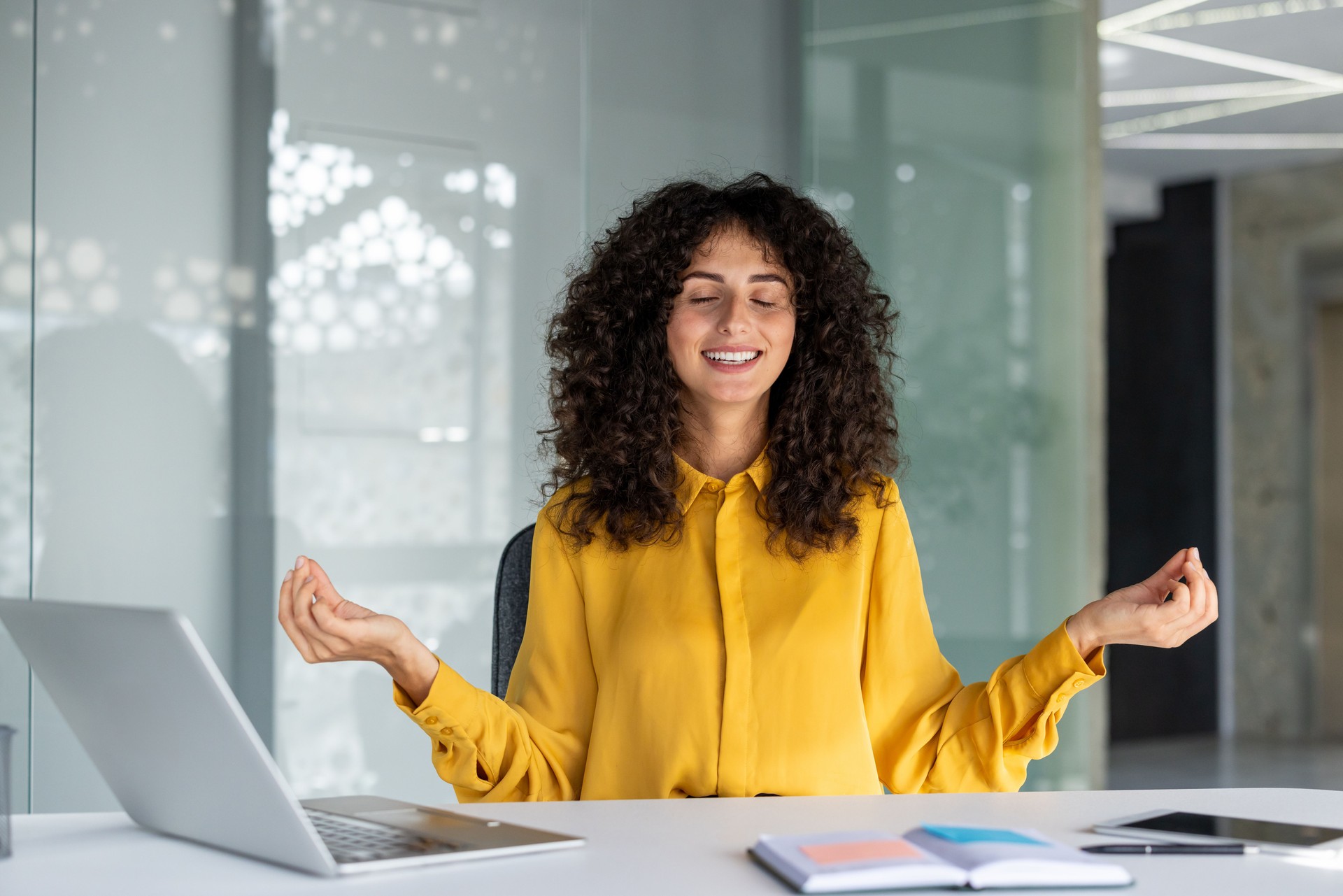 Woman practicing meditation at modern office desk with laptop and smartphone, promoting mindfulness at work. She is smiling and relaxed, wearing yellow blouse, peaceful atmosphere.